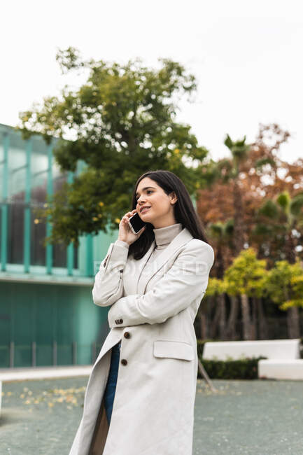 Angolo basso di felice imprenditrice parlare su smartphone e in piedi in strada mentre discuteva di progetto aziendale e guardando altrove — Foto stock