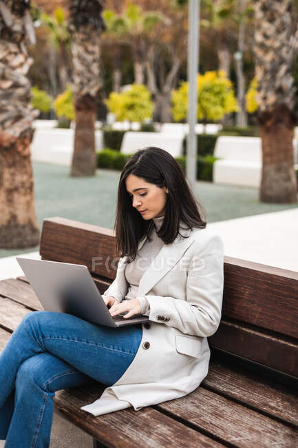 Серйозна жінка-підприємець сидить на лавці в міському парку і друкує на ноутбуці, працюючи віддалено на бізнес-проекті. — стокове фото