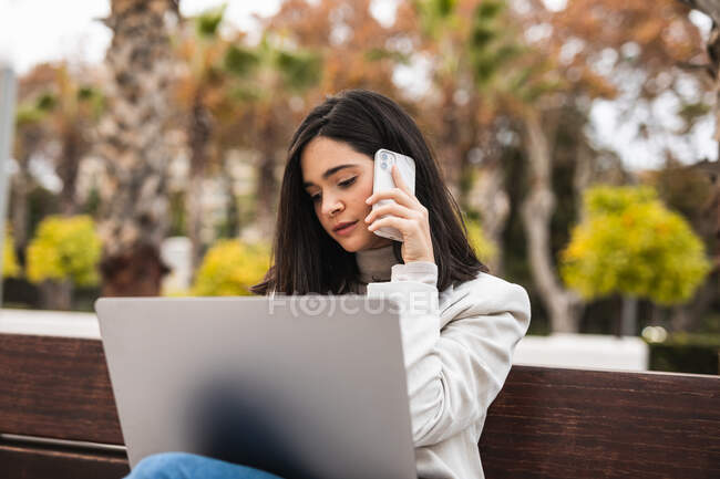 Сконцентрована бізнес-жінка сидить на лавці з ноутбуком і розмовляє смартфоном під час віддаленої роботи в міському парку. — стокове фото