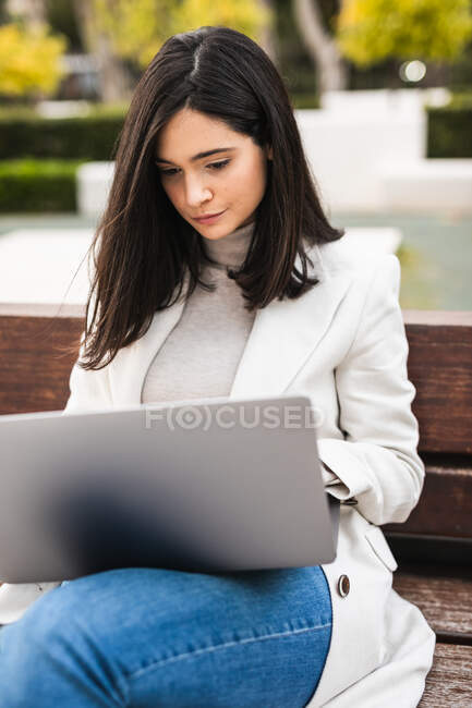 Mujer emprendedora seria sentada en el banco en el parque urbano y escribiendo en el portátil mientras trabaja remotamente en el proyecto de negocios - foto de stock
