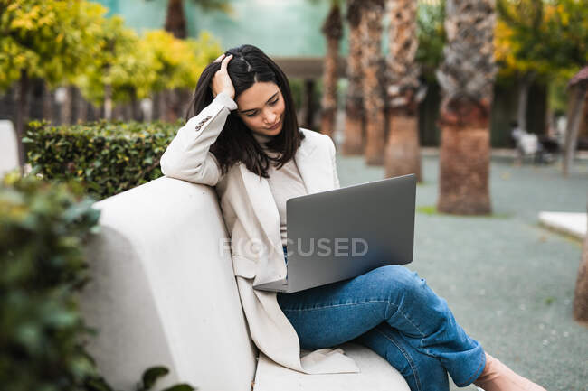 Pensive жінка-підприємець використовує ноутбук, сидячи на лавці в міському парку і працюючи в Інтернеті над проектом — стокове фото