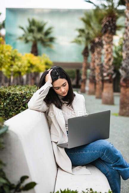 Pensive female entrepreneur using laptop while sitting on bench in city park and working online on project — Stock Photo