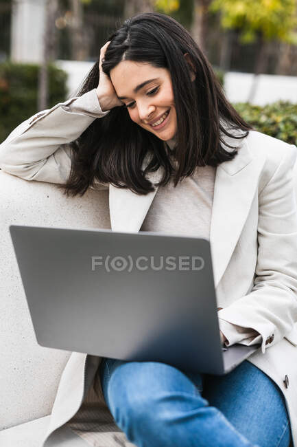 Femme entrepreneure joyeuse utilisant un ordinateur portable tout en étant assise sur un banc dans le parc de la ville et travaillant en ligne sur le projet — Photo de stock