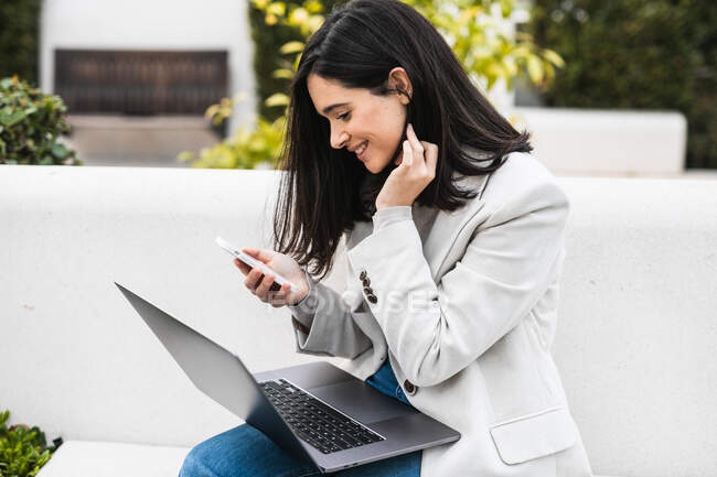 Щаслива бізнес-леді сидить на лавці з ноутбуком і переглядає смартфон під час віддаленої роботи в міському парку — стокове фото