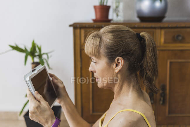 Доросла жінка в повсякденному одязі дивитися відео на сучасному планшеті у світлій вітальні — стокове фото
