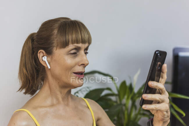 Веселая взрослая женщина в наушниках с видеозвонком через современный мобильный телефон в светлой гостиной — стоковое фото