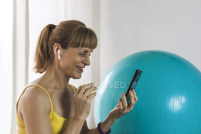 Mulher adulta alegre em fones de ouvido com videochamada através do telefone móvel moderno na sala de estar leve — Fotografia de Stock