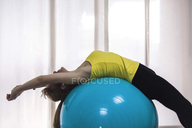 Vista lateral fêmea ajuste anônimo em activewear alongamento de volta músculos em grande bola de ioga no centro de fitness luz — Fotografia de Stock