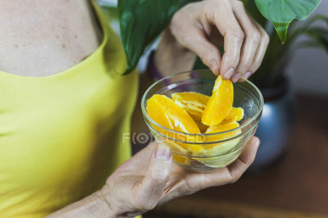 Вид збоку для дорослих жінка слухає музику через навушники і насолоджується соковитим свіжим апельсиновим сегментом у скляній мисці — стокове фото