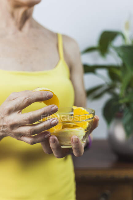 Cosecha hembra adulta escuchando música a través de auriculares y disfrutando de un jugoso segmento de naranja fresca en un tazón de vidrio - foto de stock