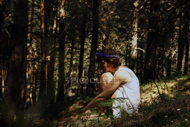 Side view of romantic young lady with short hair in summer dress and floral wreath embracing knees while sitting on grass in lush forest — Stock Photo