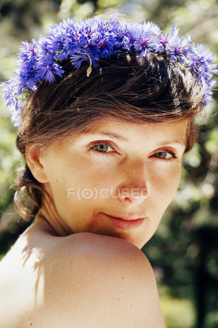 Peaceful adult female with bare shoulder and floral wreath on head standing looking at camera on sunny day in forest — Stock Photo