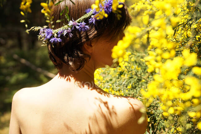 Vista posterior de hembra desnuda adulta tranquila con corona floral en la cabeza descansando en el jardín cerca del árbol floreciente con flores amarillas en el día soleado - foto de stock