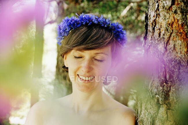 Peaceful delighted adult female with bare shoulder and floral wreath on head standing near tree with eyes closed on sunny day in forest — Stock Photo