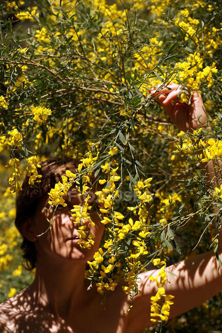 Femme nue adulte calme aux yeux fermés se reposant dans le jardin près d'un arbre en fleurs jaunes par une journée ensoleillée — Photo de stock