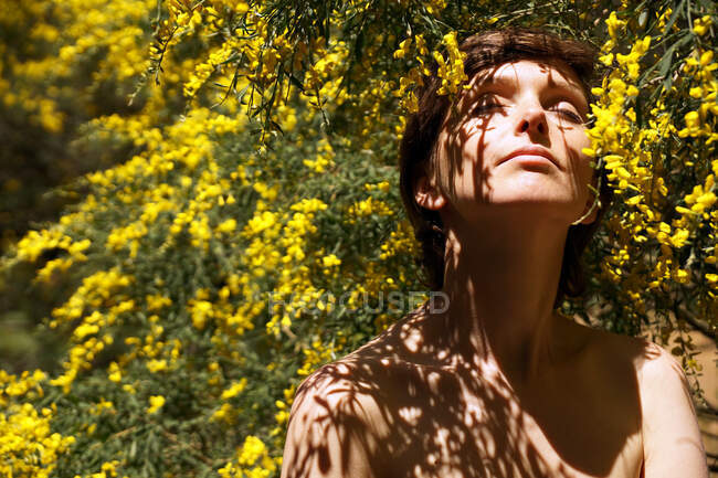 Спокойная взрослая обнаженная женщина с закрытыми глазами отдыхает в саду рядом с цветущим деревом с желтыми цветами в солнечный день — стоковое фото
