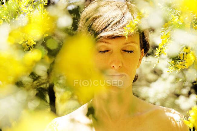 Спокойная взрослая обнаженная женщина с закрытыми глазами отдыхает в саду рядом с цветущим деревом с желтыми цветами в солнечный день — стоковое фото