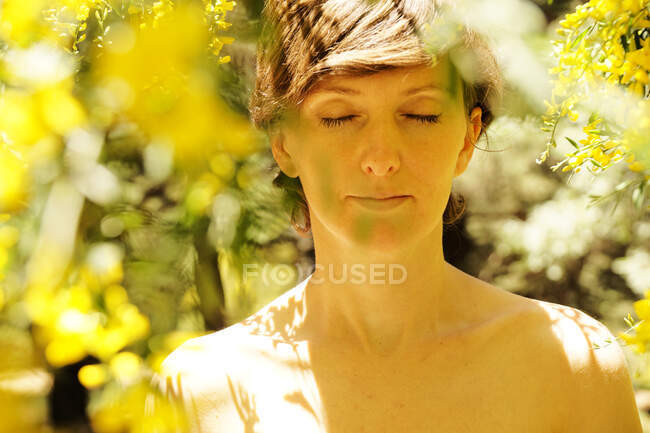 Calmo adulto fêmea nua com olhos fechados descansando no jardim perto da árvore florescente com flores amarelas no dia ensolarado — Fotografia de Stock