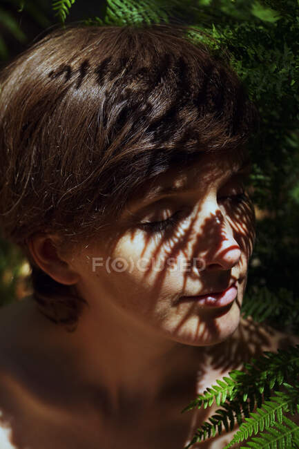 Atractivo joven mujer desnuda con el pelo oscuro sentado cerca de helecho arbusto en exuberante bosque tropical con los ojos cerrados en el día soleado - foto de stock