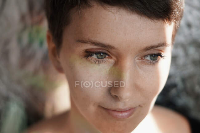 Cosecha femenina dama adulta reflexiva con maquillaje natural y pelo corto sonriendo y mirando hacia otro lado soñando a la luz del sol - foto de stock