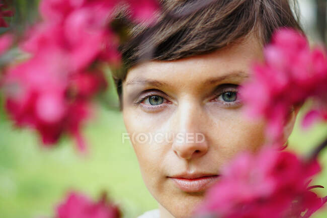 Задумчивый урожай взрослой женщины с короткими волосами воссоздания в зеленом саду рядом с яркими цветущими цветами и смотреть в камеру — стоковое фото