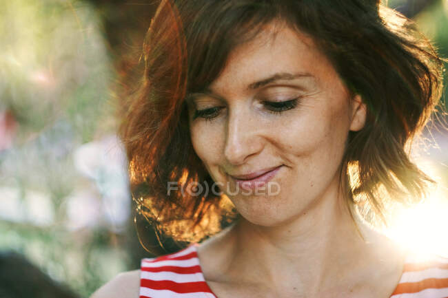 Crop thoughtful happy adult female resting in nature and looking down pensively under bright sunlight during golden hour — Stock Photo