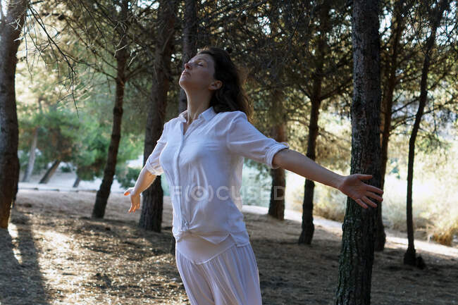 Peaceful adult female in casual clothes meditating in forest with closed eyes and outstretched arms in sunny morning — Stock Photo