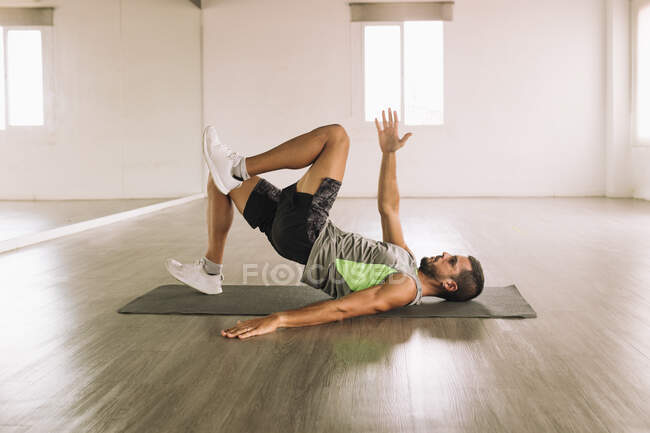 Вид сбоку на мускулистого молодого бородатого спортсмена, лежащего на коврике возле зеркала и занимающегося маршем Glute Bridge, тренируясь в одиночестве в просторной легкой студии — стоковое фото