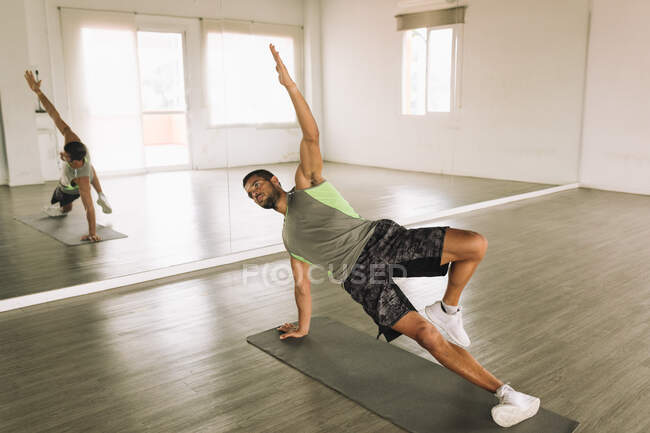 Full body of young focused fit determined sportsman in activewear performing High Side Plank with leg raises exercise during workout in studio near big wall mirror — Stock Photo