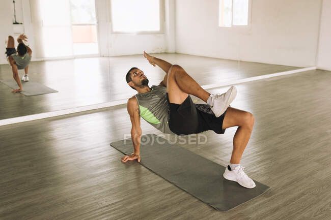 Вид сбоку на мускулистого молодого бородатого спортсмена, лежащего на коврике возле зеркала и занимающегося маршем Glute Bridge, тренируясь в одиночестве в просторной легкой студии — стоковое фото