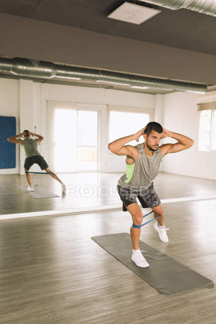 Konzentrierter junger athletischer Typ in Sportbekleidung, der Side Lunges Übungen mit Gummiband macht, während er allein im geräumigen Fitnessstudio mit Spiegel trainiert — Stockfoto