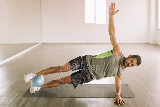 Vista lateral de determinado deportista joven muscular en ropa deportiva haciendo ejercicio Leg Crunch con balón de medicina mientras está acostado en la estera durante el entrenamiento en el estudio de luz - foto de stock