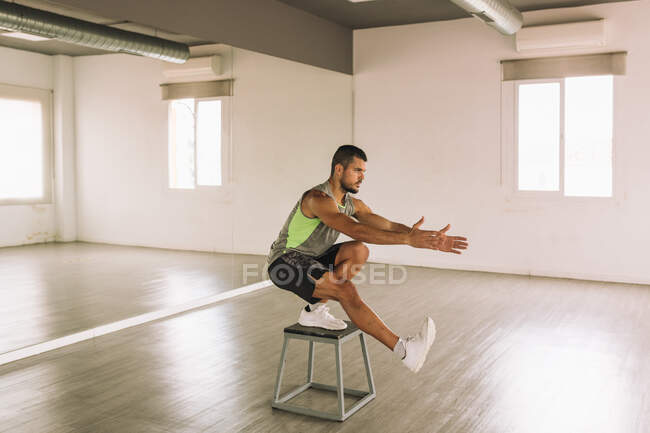 Vista lateral de un hombre atlético joven en forma seria en ropa deportiva haciendo ejercicio de sentadilla de una pierna en la plataforma de paso mientras se entrena en el estudio - foto de stock
