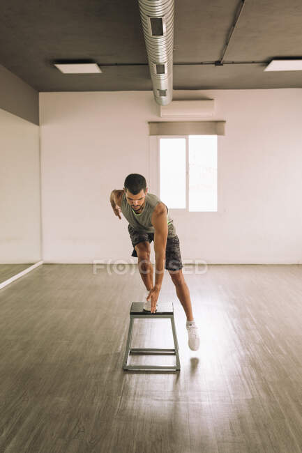 Longitud completa de los jóvenes deportistas musculosos enfocados practicando Squat Pop Over ejercicio en la plataforma de paso mientras se ejercita solo en el estudio de luz - foto de stock