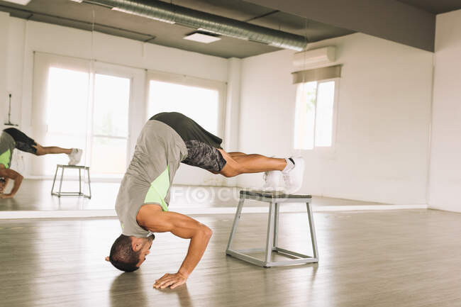Vista lateral do jovem ginasta masculino muscular fazendo Pike Push Ups com pernas na plataforma de passo durante o treinamento em estúdio perto do espelho — Fotografia de Stock