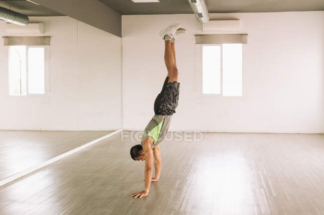 Side view of young strong fit guy in sportswear standing on hands while training alone in spacious light studio — Stock Photo