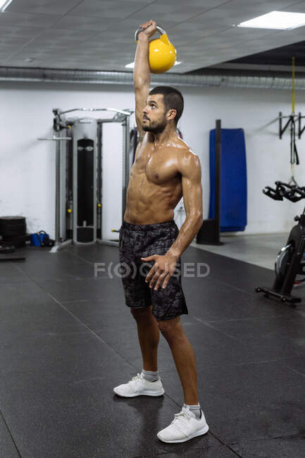 Vista lateral de determinado joven musculoso deportista sin camisa levantando pesadas pesas pesas pesas mientras entrena bíceps en el gimnasio moderno - foto de stock