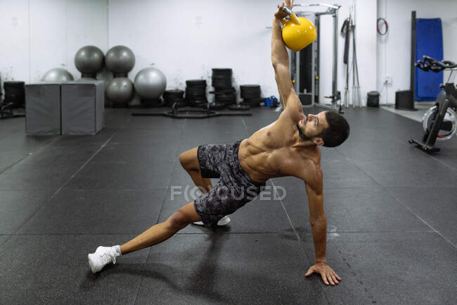 Poderoso muscular jovem atleta masculino com tronco nu em pé na prancha lateral e levantando kettlebell pesado durante o treino no ginásio — Fotografia de Stock