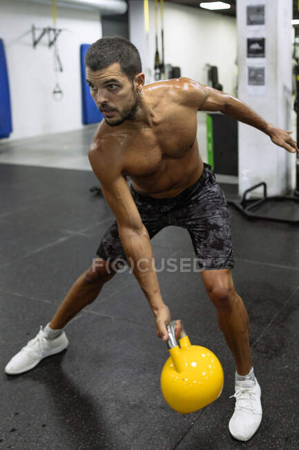 Сильний мускулистий молодий спортсмен з голим тулубом стоїть на схилі планки і піднімає важкий кетлебел під час тренувань у спортзалі. — стокове фото