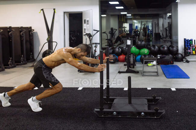 Vista lateral de un joven deportista sudado sin camisa decidido haciendo ejercicio de empuje de trineo con equipo pesado en el gimnasio moderno - foto de stock