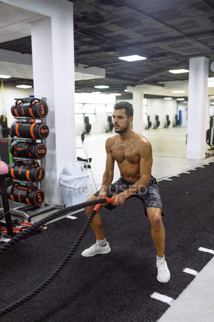 Мускулистый молодой спортсмен без рубашки тренируется с боевыми веревками во время интенсивной тренировки в современном тренажерном зале — стоковое фото