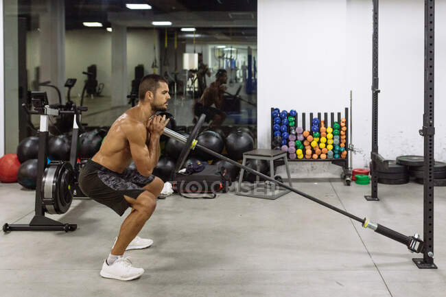 Vista lateral de un joven deportista muscular fuerte concentrado con barra de elevación del torso desnuda mientras entrena duro en el gimnasio contemporáneo - foto de stock