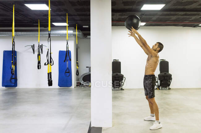Вид сбоку на выносливого молодого спортсмена с обнаженным туловищем, бросающего мячик тяжелой медицины во время интенсивной тренировки в современном спортзале — стоковое фото