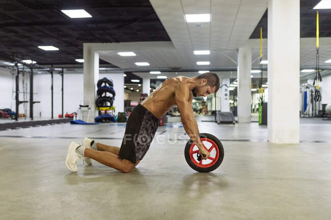 Visão lateral do jovem musculoso forte suado usando rolo de roda enquanto faz exercício abdominais no clube esportivo contemporâneo — Fotografia de Stock