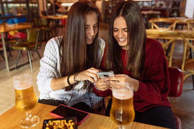 Молода весела лесбійська пара сидить за столом з келихами пива в кафе і використовує смартфон, проводячи вихідні разом — стокове фото