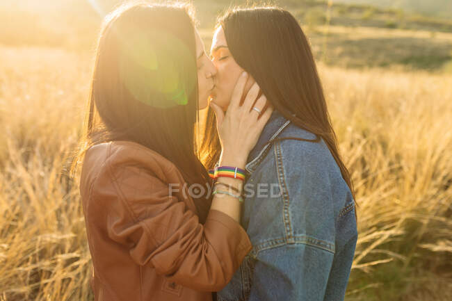 Vista lateral de joven pareja lesbiana de pie en el campo y besándose tiernamente con los ojos cerrados - foto de stock