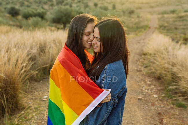 Vue latérale en angle élevé d'un couple de lesbiennes douces enveloppées dans un drapeau arc-en-ciel LGBT blottissant sur une route sablonneuse dans la nature avec les yeux fermés et le sourire — Photo de stock