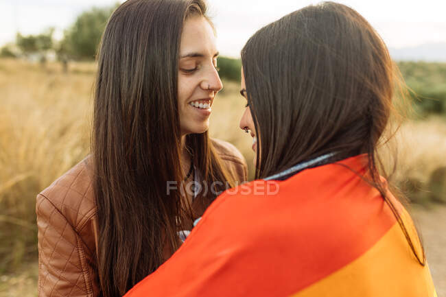 Побічний вид на лагідну пару лесбійських жінок, загорнутих в ЛГБТ райдуга прапор пригортаючись до піщаної дороги в природі з закритими очима і посміхаючись — стокове фото
