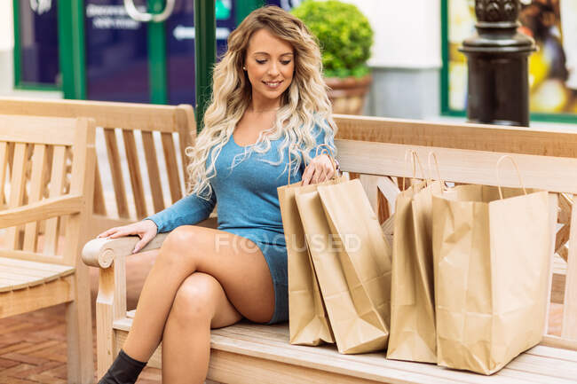 Mujer feliz sentado en el banco y mirando las mercancías en bolsas de compras en la ciudad - foto de stock