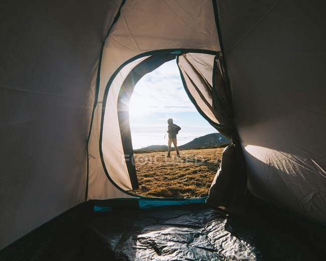 Обратный вид на неузнаваемого путешественника в теплой одежде и рюкзаке, стоящего на травянистой местности возле палатки во время похода в высокогорье в солнечное утро — стоковое фото